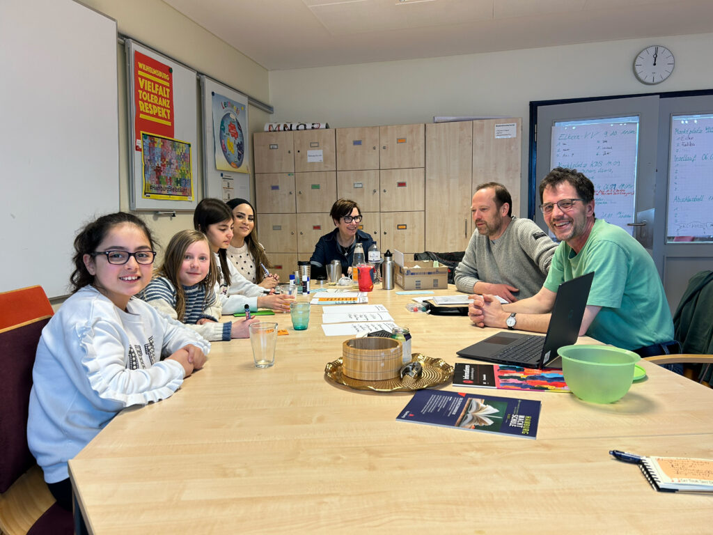 Die Arbeitsgruppe Homepage: Schülerinnen und Lehrkräfte bei der gemeinsamen Arbeit im Menschenzimmer an einem großen Tisch mit Moderationskarten, Tablet und Laptop