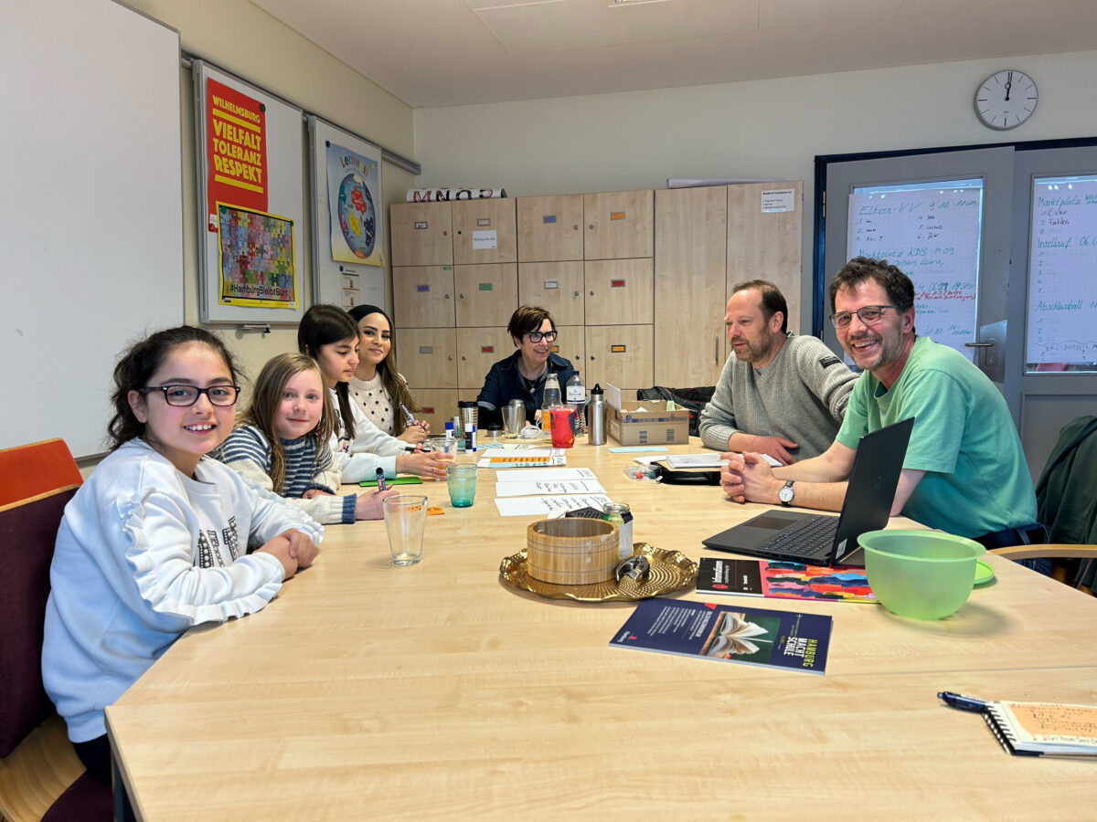Die Arbeitsgruppe Homepage: Schülerinnen und Lehrkräfte bei der gemeinsamen Arbeit im Menschenzimmer an einem großen Tisch mit Moderationskarten, Tablet und Laptop