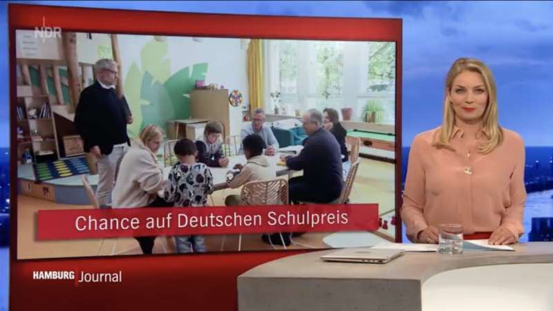 Eine Moderatorin kündigt den Bericht über den Besuch der Schulpreisjury an