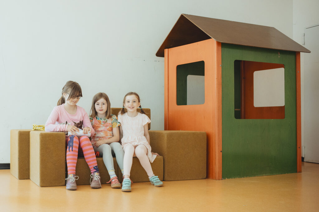 Drei Kinder sitzen auf der Bühne der Pausenhalle auf Kisten vor einem großen Spielhaus.