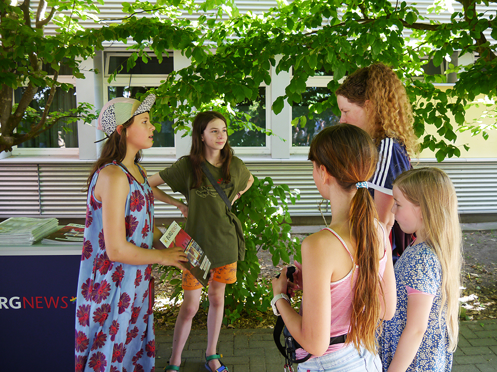 Am Stand der Schüler*innenzeitung wird die neueste Ausgabe der  BurgNews angeboten. Schülerinnen im Gespräch.